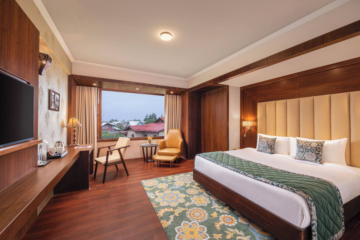 A hotel room with a large bed, a desk with a flat-screen TV, a chair, a small table with a lamp, and a view of the outside.