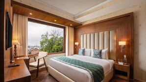 A hotel room with a large bed, a desk with a chair, a TV, and a window with a view of buildings and trees. - WelcomHeritage Grand Srinagar (Srinagar)