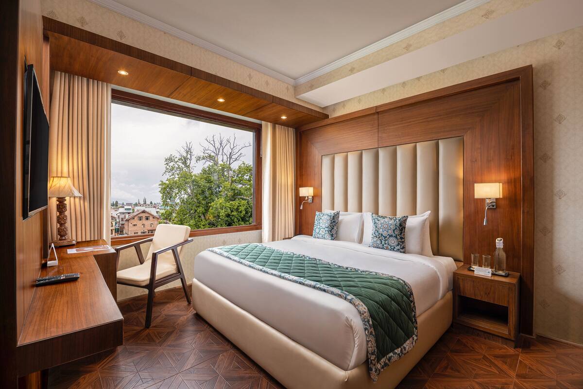 A hotel room with a large bed, a desk with a chair, a TV, and a window with a view of buildings and trees.