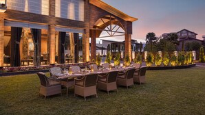 An outdoor dining area with wicker furniture and a large table set for a meal, surrounded by a well-maintained lawn and a building with large windows and a wooden facade. - WelcomHeritage Grand Srinagar (Srinagar)