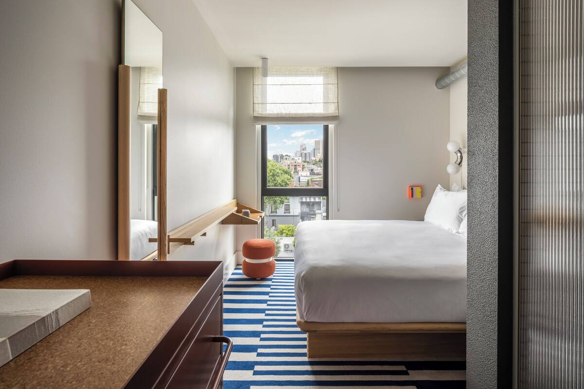 Una habitación de hotel moderna con una cama grande, una mesita de noche de madera, un espejo y vistas de la ciudad desde la ventana.