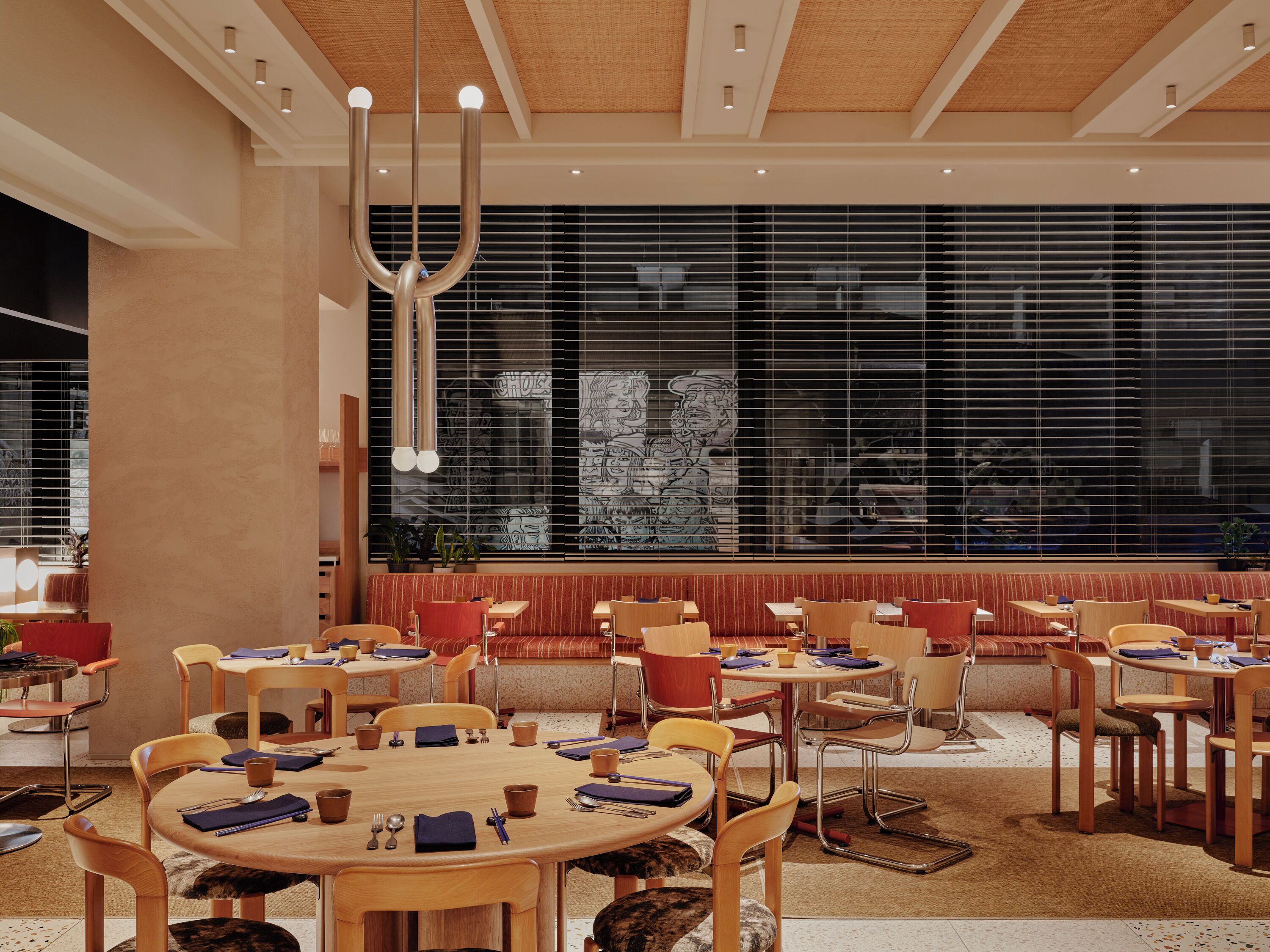 A modern dining area with round tables and chairs, large windows with blinds, and a ceiling with recessed lighting.