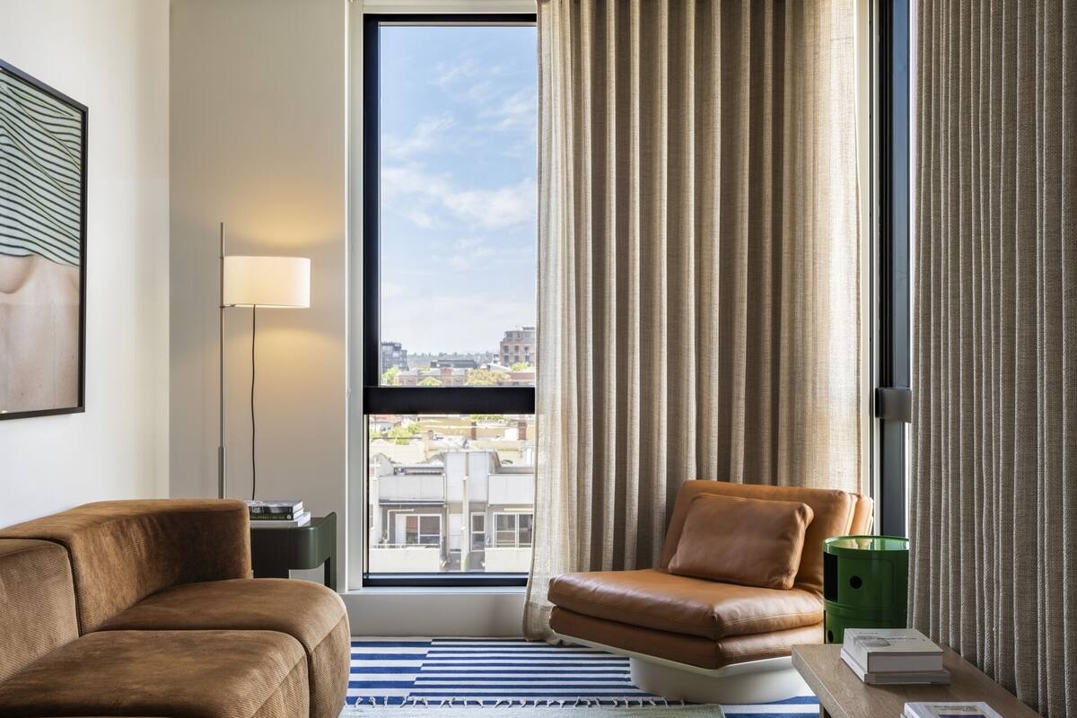 Una habitación de hotel moderna con un ventanal grande, un sillón de cuero marrón, un tapete azul a rayas y una pequeña mesa con libros.