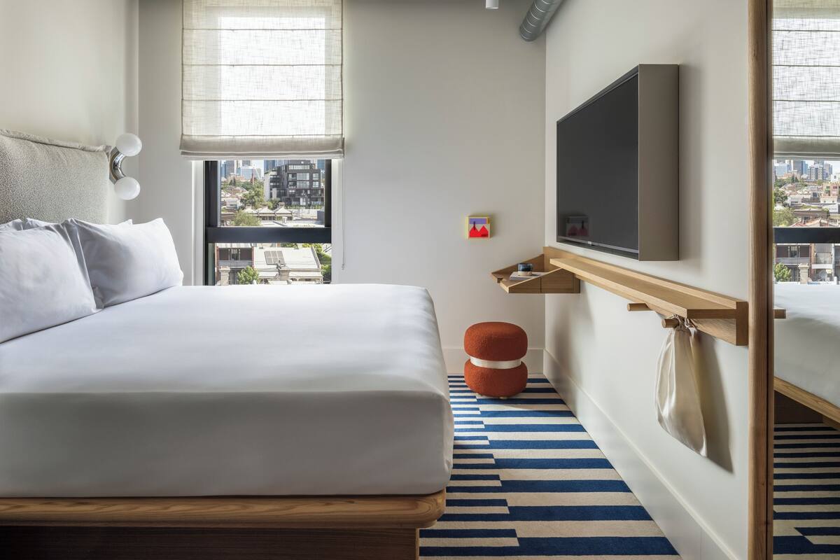 Una habitación de hotel moderna con una cama grande, un televisor montado en la pared, un ventanal con vistas a la ciudad y una alfombra a rayas.
