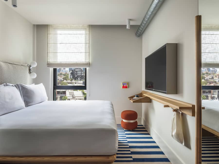 Une chambre d’hôtel moderne avec un grand lit, un téléviseur fixé au mur, une fenêtre donnant sur la ville et un tapis à rayures.
