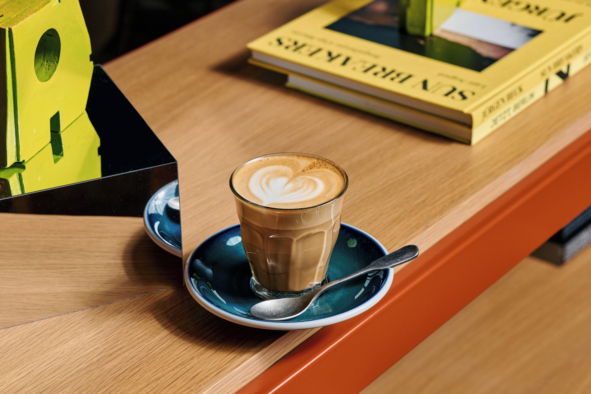 Une tasse de café ornée d’un motif en latte art, posée sur une soucoupe, avec une cuillère à proximité et un livre intitulé « SLIN THIRTEEN » sur une table en bois.