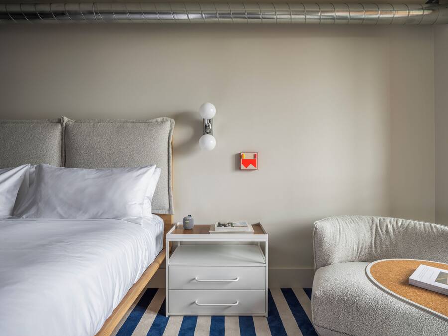 A neatly arranged hotel room with a bed, a bedside table with drawers, a lamp, and a chair with a small table.