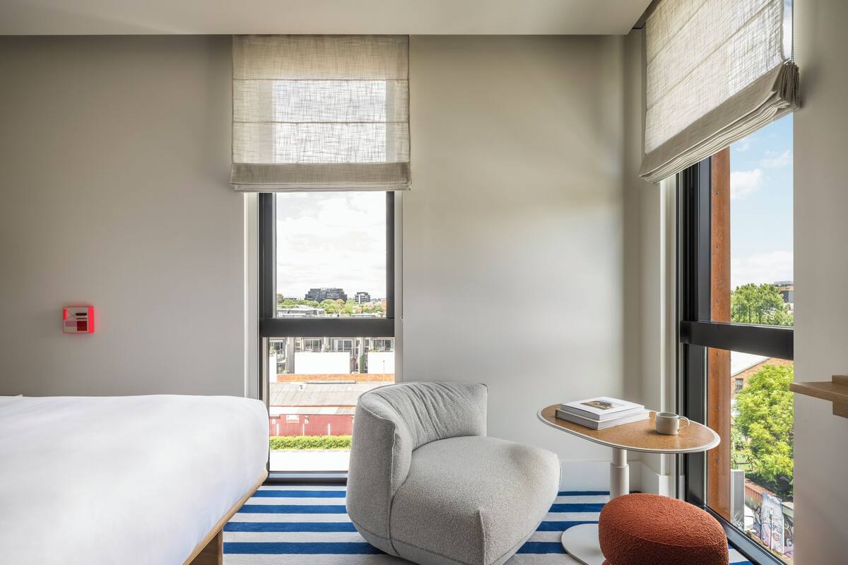 Habitación de hotel con cama, un sillón gris, una pequeña mesa redonda con un libro y una taza, y un ventanal con vistas a edificios y árboles.