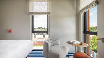 A hotel room with a bed, a gray armchair, a small round table with a book and a cup, and a large window with a view of buildings and trees.
