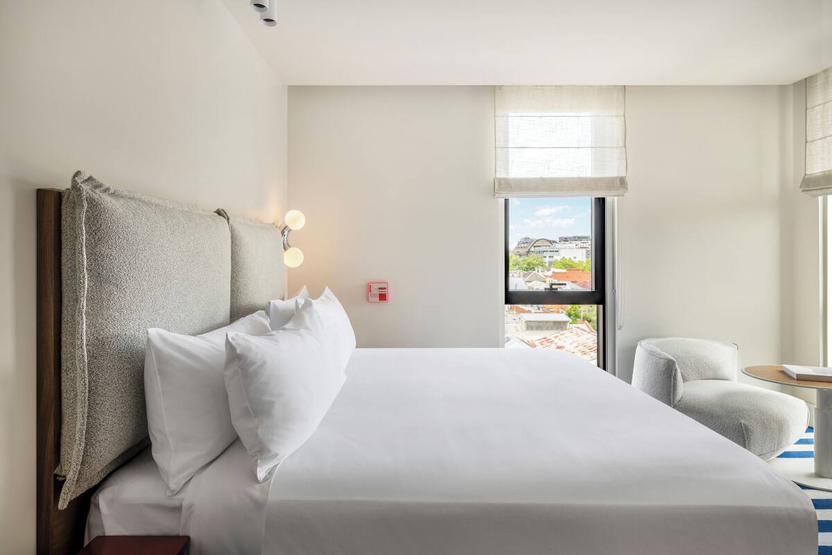 Habitación de hotel con una cama grande, una silla, un ventanal con vistas a edificios y una lámpara fijada a la pared.