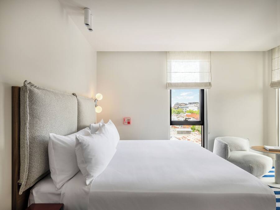 A hotel room with a large bed, a chair, a window with a view of buildings, and a wall-mounted light fixture.