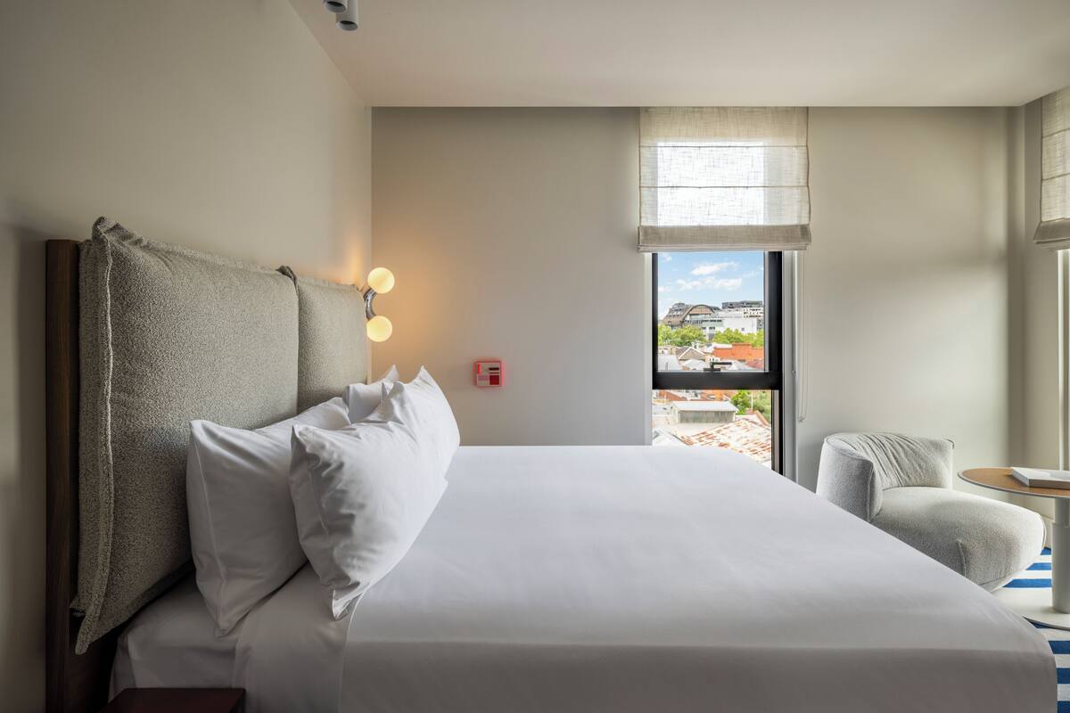 Habitación de hotel con una cama grande, una silla, una mesita y una ventana con vistas a edificios.