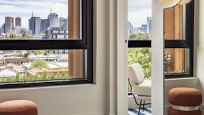 Una habitación moderna con ventanas amplias que ofrecen vistas de la ciudad, una silla blanca y dos otomanas redondas.