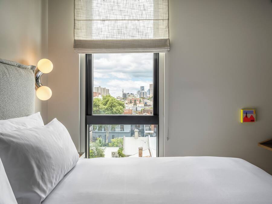 Habitación de hotel con un ventanal grande, una cama con ropa de cama blanca, una lámpara de pared y vistas a la ciudad.