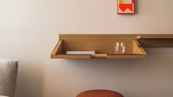 A room with a wooden shelf holding books and glasses, a red ottoman, and a striped rug.
