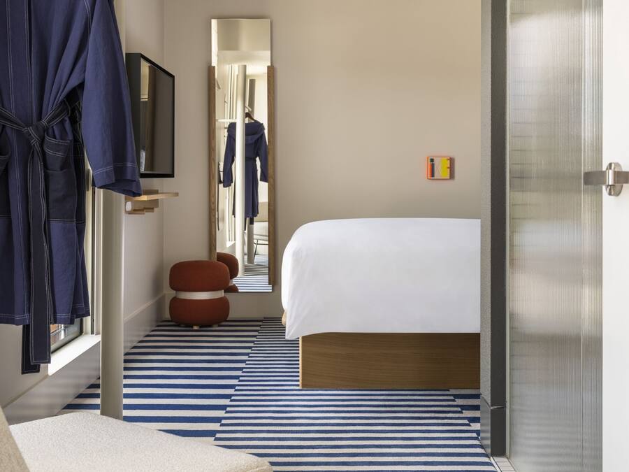Quarto de hotel com cama, um espelho grande, roupão pendurado, um banquinho redondo e piso listrado.