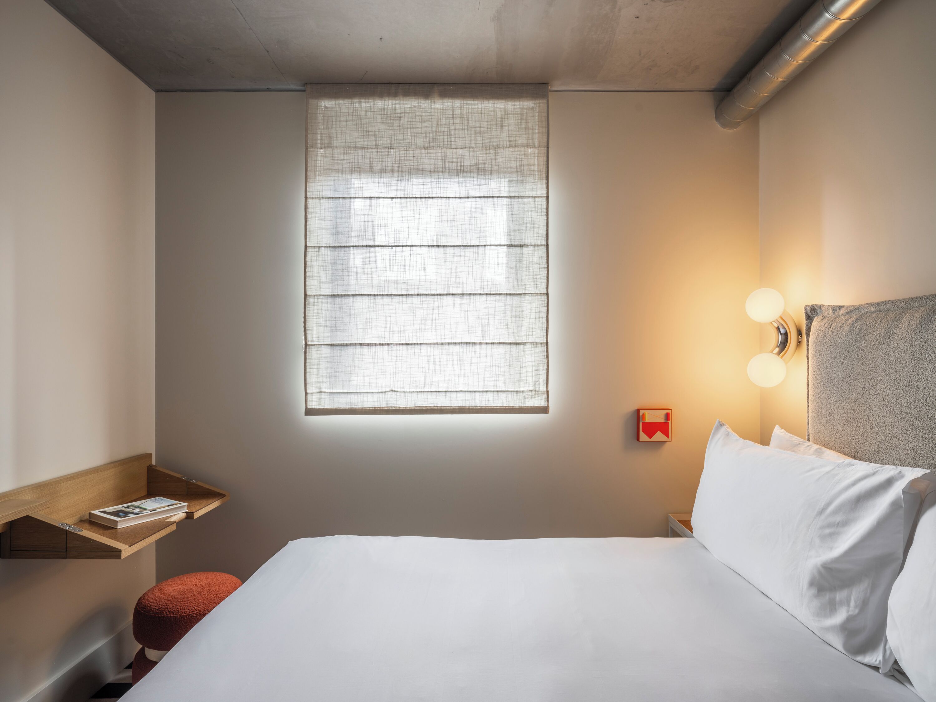 Een slaapkamer met een bed, een raam, een nachtkastje met een boek en een wandlamp.