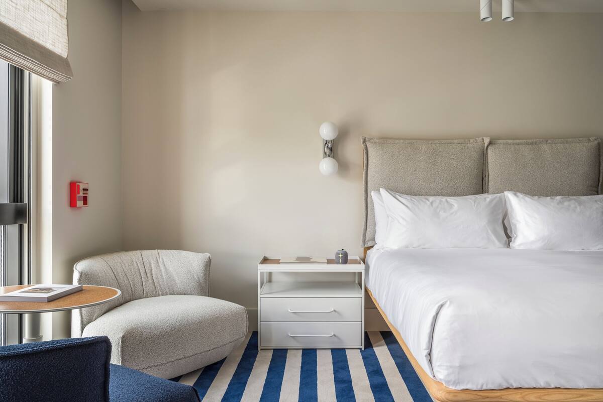 Una habitación de hotel moderna con una cama grande, una pequeña zona de estar, una mesita de noche y un ventanal con persianas.