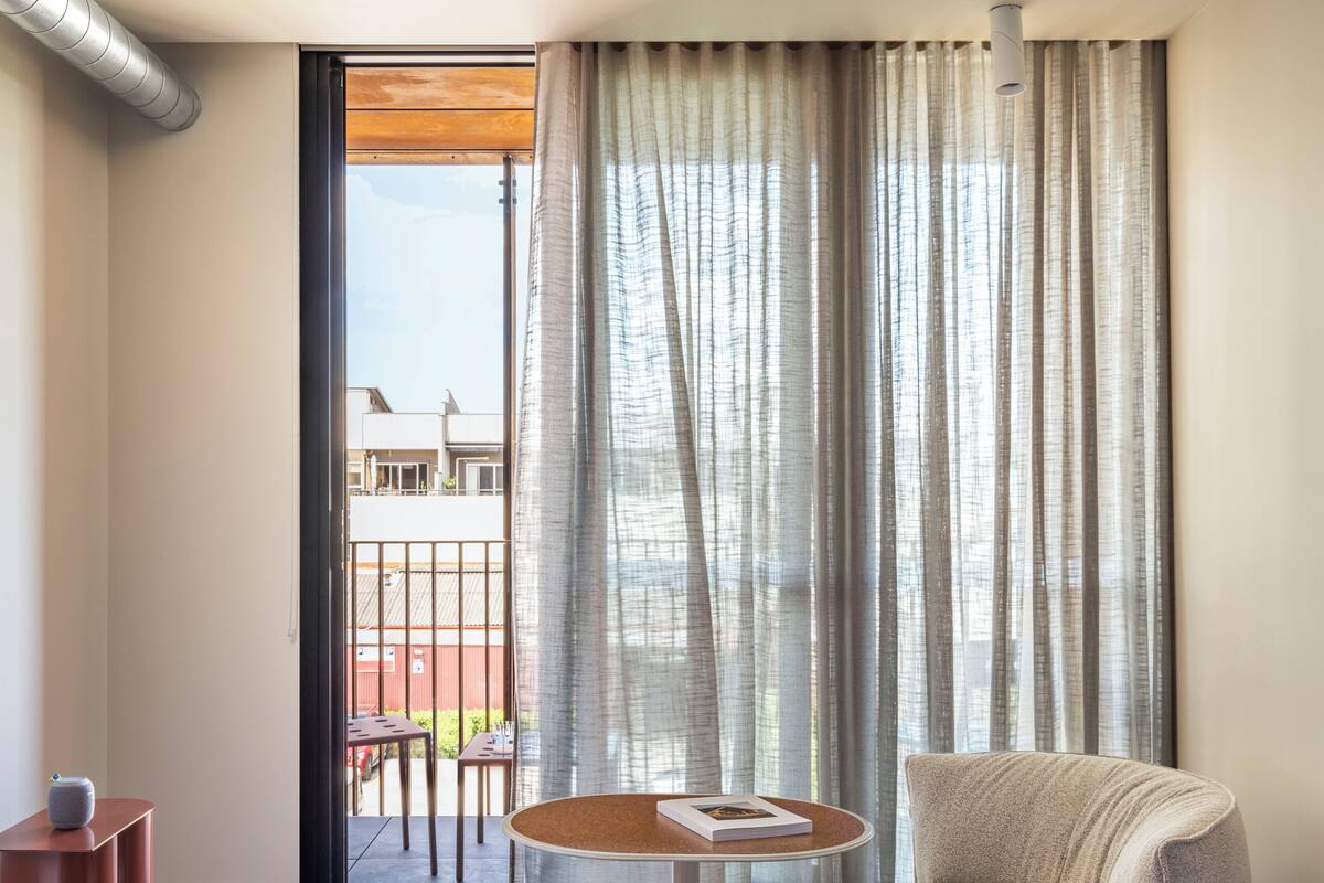 Una habitación de hotel moderna con un ventanal grande, una mesa redonda con un libro, un sillón color beige y un tapete a rayas.