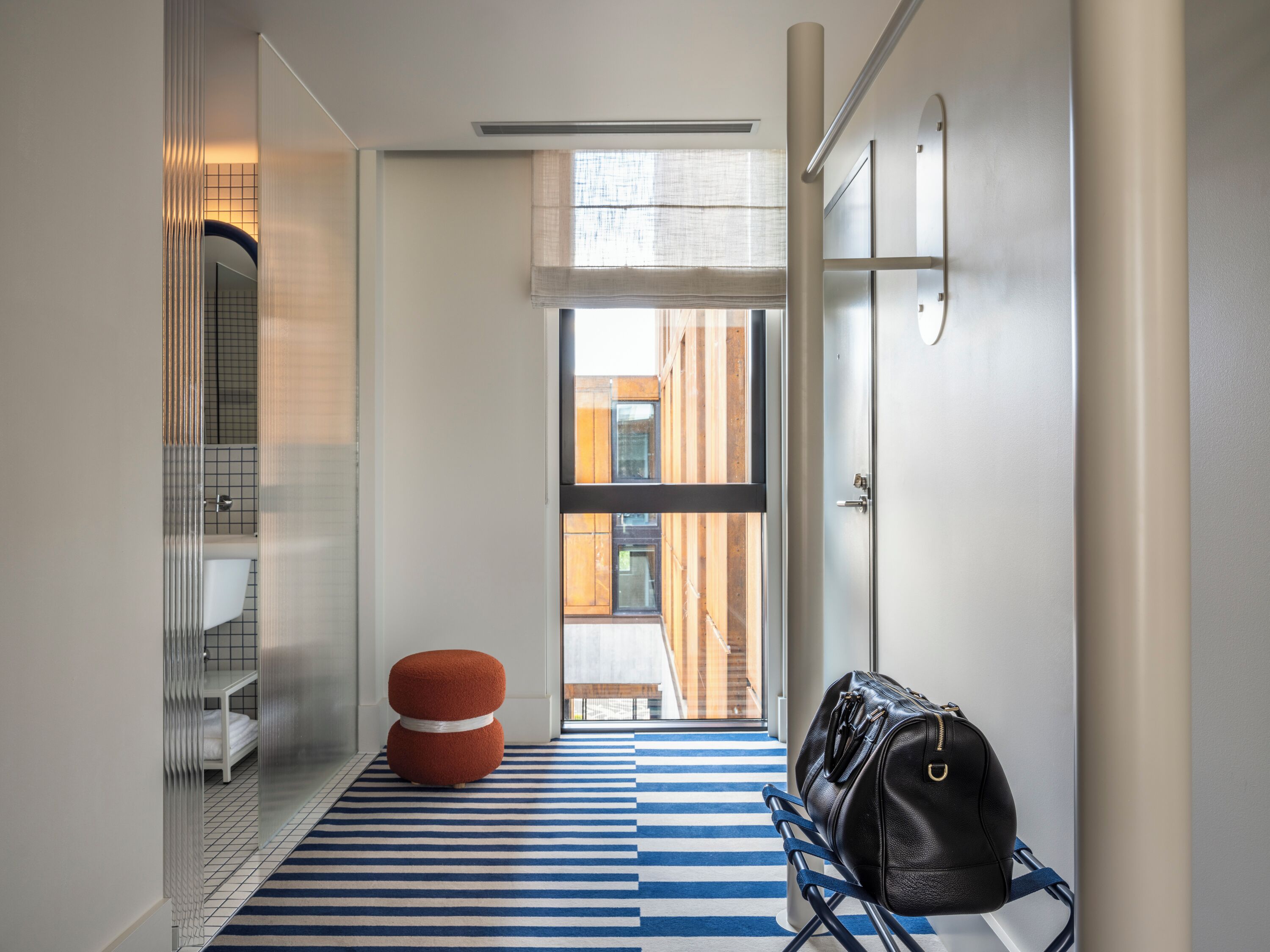 Ein Hotelkorridor mit gestreiftem Boden, einem roten Hocker und einer schwarzen Tasche auf einem Ständer.