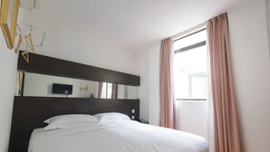 Una cama bien tendida en una habitación de hotel, con un espejo grande arriba, una ventana con cortinas y suelo de madera.