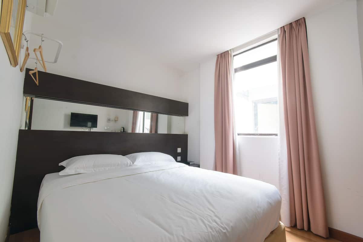 A neatly made bed in a hotel room with a large mirror above it, a window with curtains, and a wooden floor.