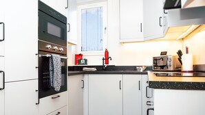 A compact kitchen with white cabinets, a black countertop, a built-in oven, a toaster, and a window with blinds. - Haywood by Central Serviced Apartments (Dundee City Council)