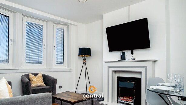 A modern living room with a fireplace, a flat-screen TV mounted above it, a sofa, a coffee table, and a dining area with a glass table and chairs. - Haywood by Central Serviced Apartments (Dundee City Council)