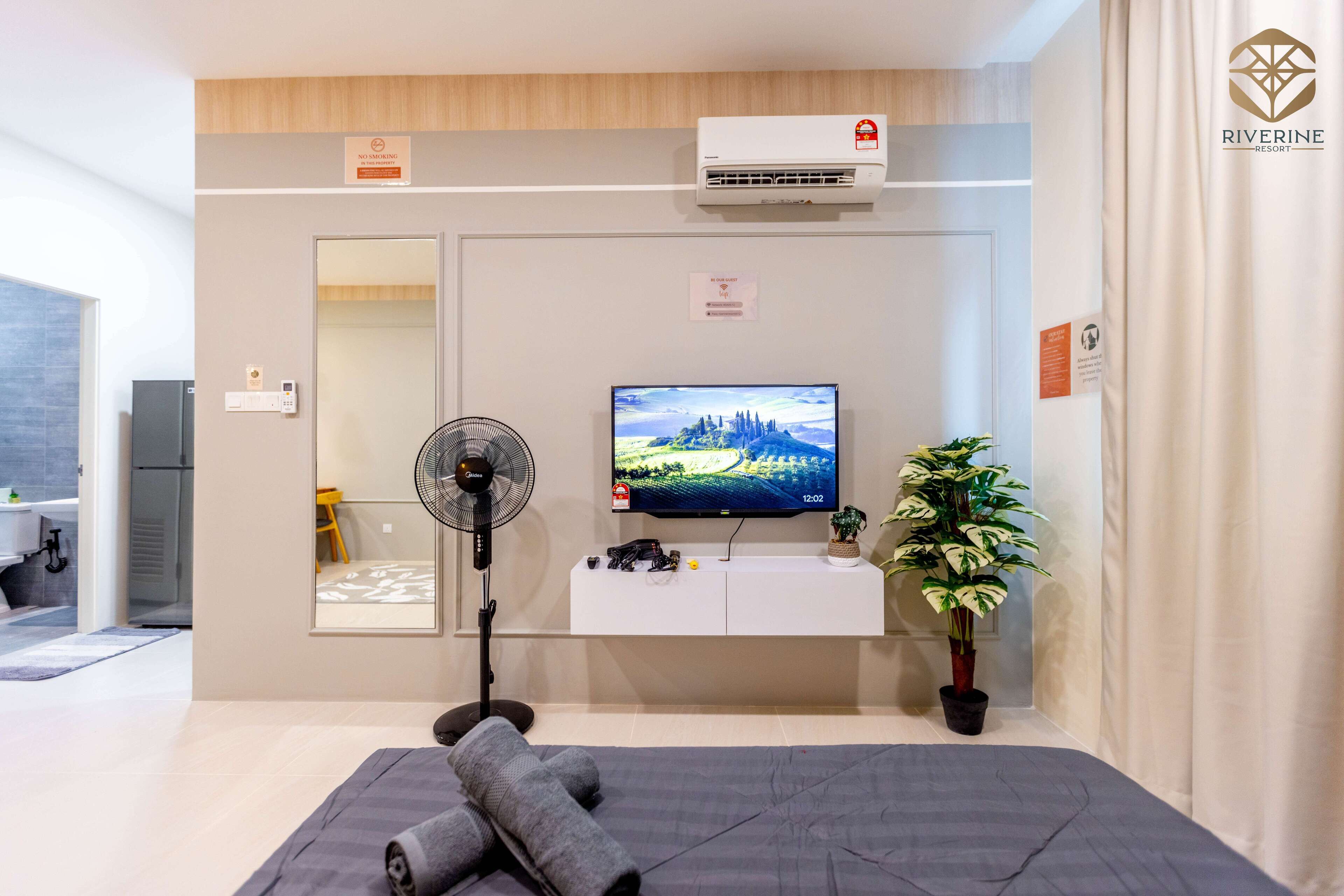 Une chambre d’hôtel moderne équipée d’un téléviseur à écran plat, d’un ventilateur sur pied, d’une plante en pot et d’un lit avec des couvertures pliées.