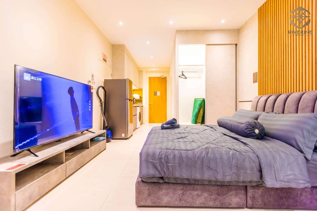 A modern hotel room with a large flat-screen TV, a bed with a grey comforter, a wooden panel wall, and a small kitchenette.