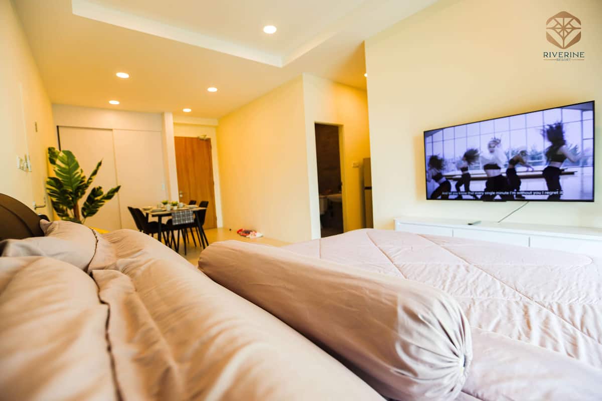 A modern hotel room with a large bed, a television on the wall, and a dining area in the background.