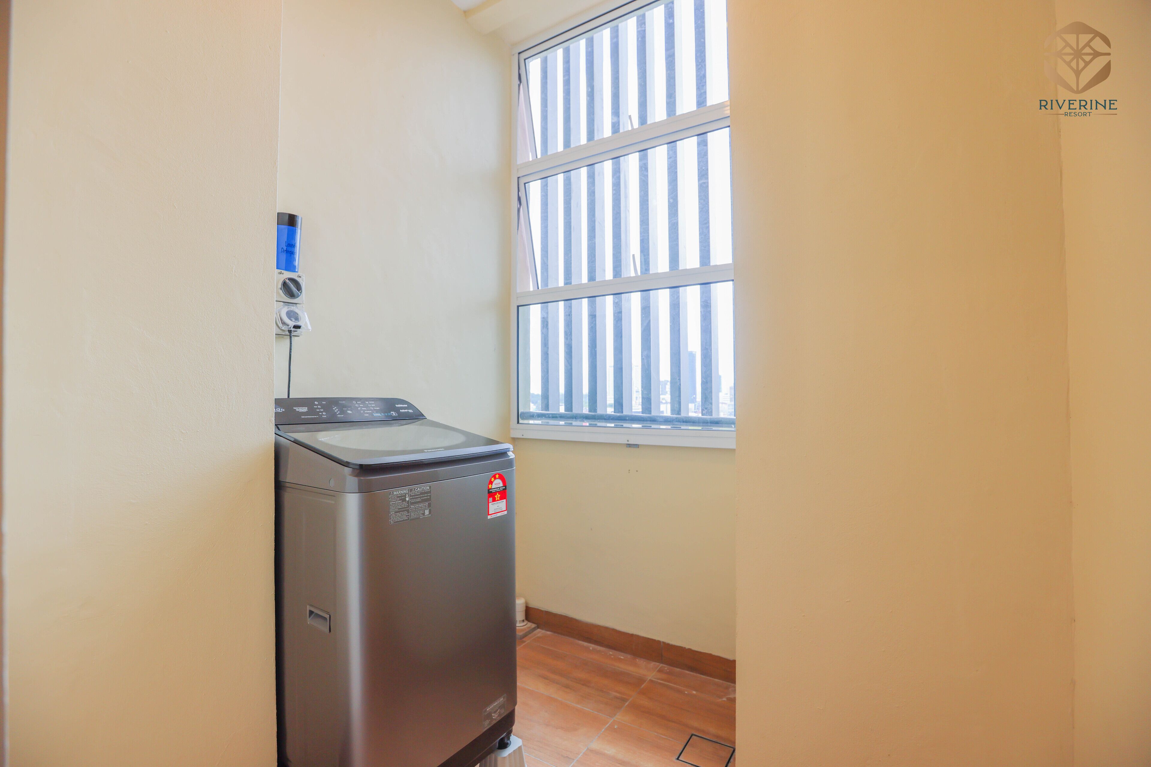 A compact room with a washing machine, a window with blinds, and a visible Riverine Resort logo.