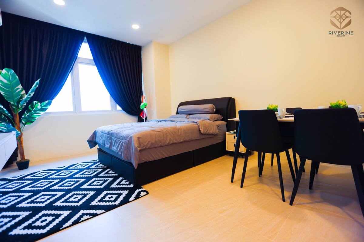 A modern hotel room with a bed, a desk with chairs, a potted plant, and a window with curtains.