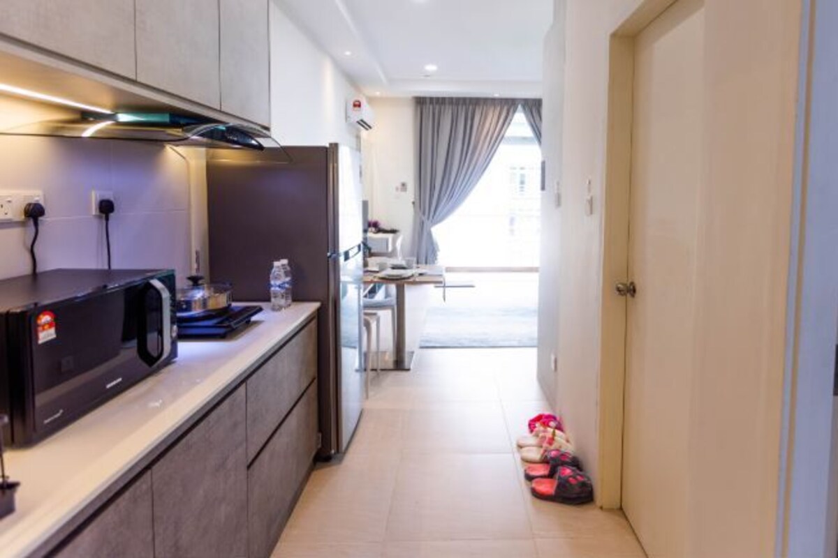 A modern kitchen with a microwave, a refrigerator, and a countertop with various items. There is a hallway leading to a room with a door and a window with curtains.