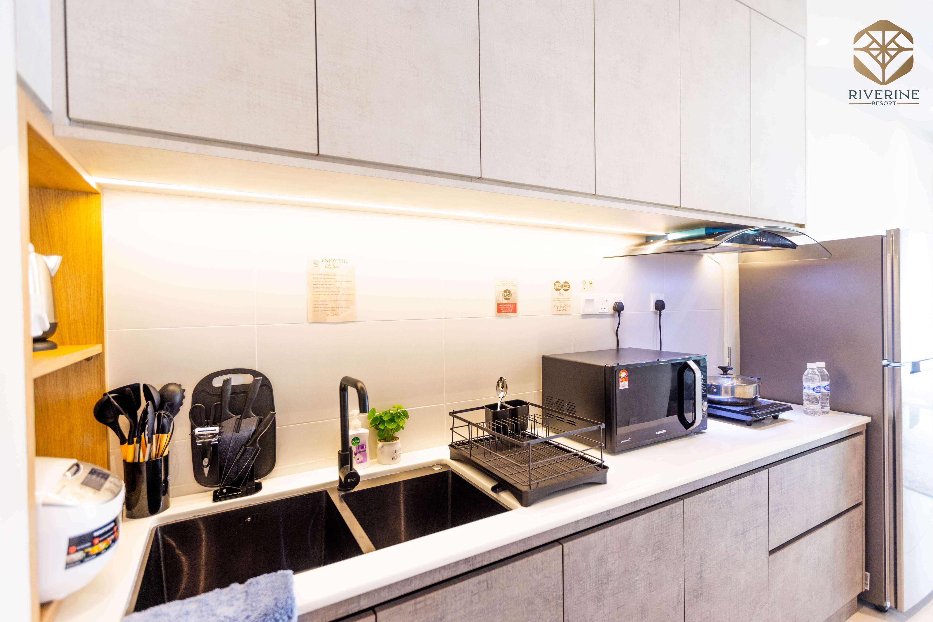 A modern kitchen with a stainless steel sink, a microwave, and a kettle. There are utensils and a small plant on the counter.