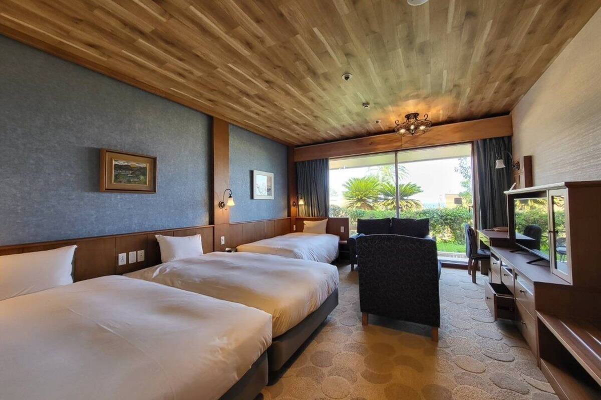 Habitació d'hotel amb dos llits, una taula, una cadira i un finestra gran amb vista a vegetació.