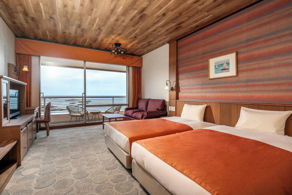 Habitació d'hotel amb dos llits, un escriptori amb televisió, una cadira, un sofà i una finestra gran amb vista al mar.