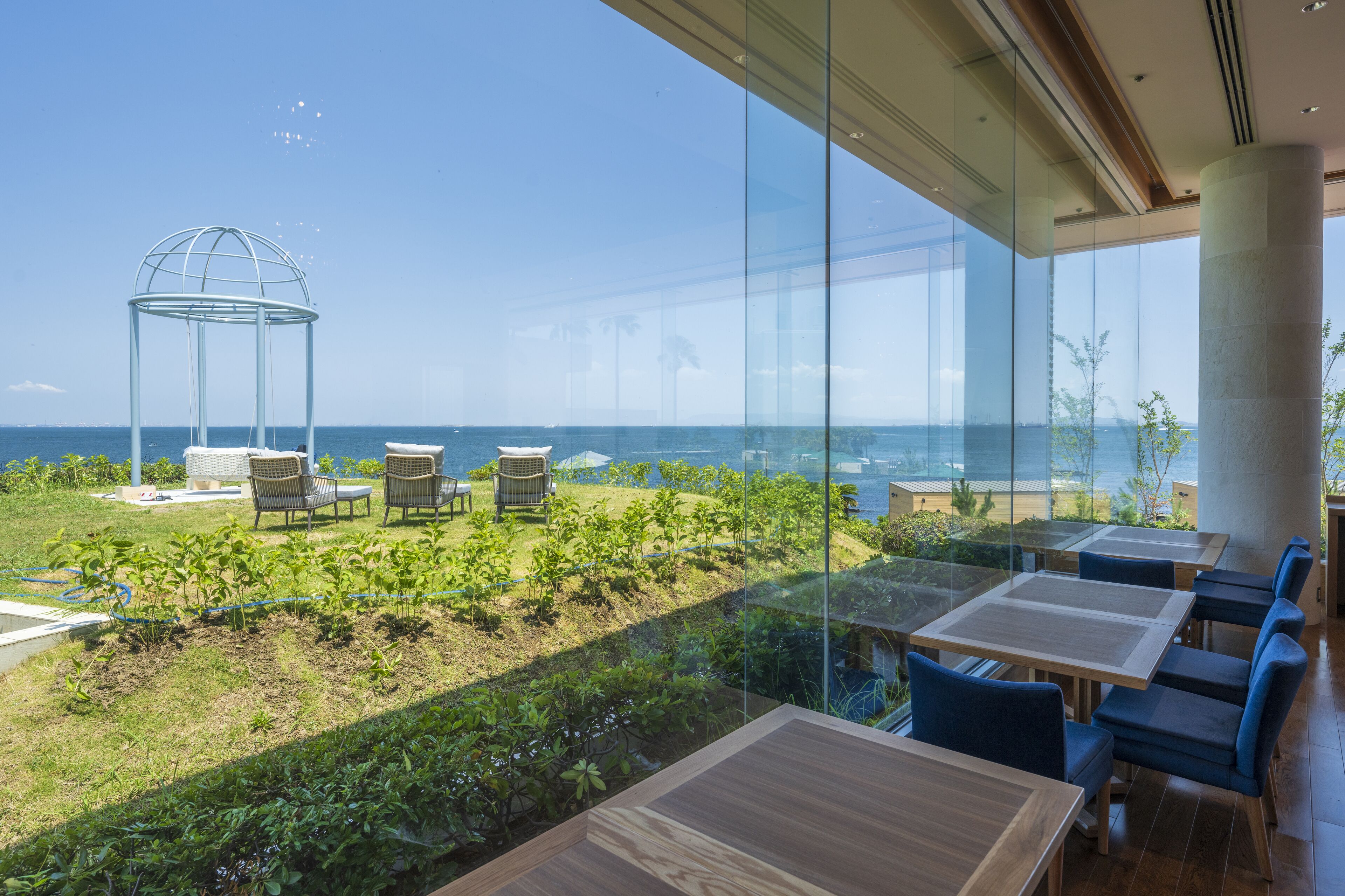 A modern outdoor dining area with glass walls, a garden, and a view of the sea.