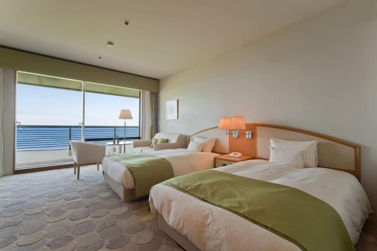 A hotel room with two beds, a sofa, a chair, a desk, and a view of the sea.