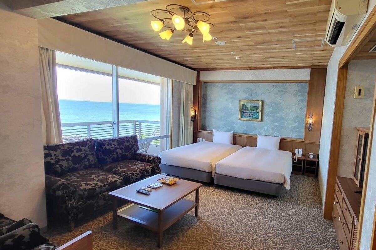 Habitació d'hotel amb una finestra gran que ofereix vista al mar, un sofà, dos llits, un sostre de fusta i una catifes amb dibuixos.