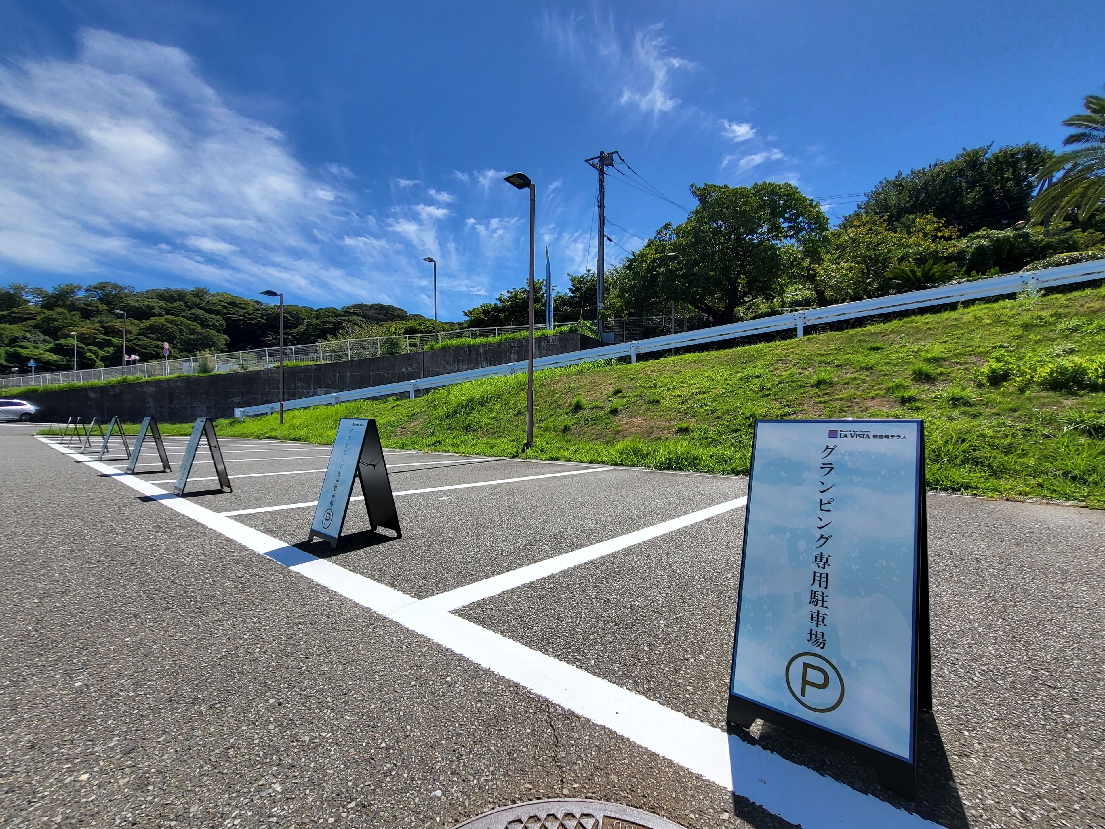 A parking area with marked spaces, a signboard, and a grassy slope with a fence.