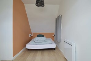 A small, single-bed room with a sloped ceiling, a radiator, and a wall-mounted light fixture. - - La Petite Baleine, Appt à 50m de la Plage - (Luc-sur-Mer)