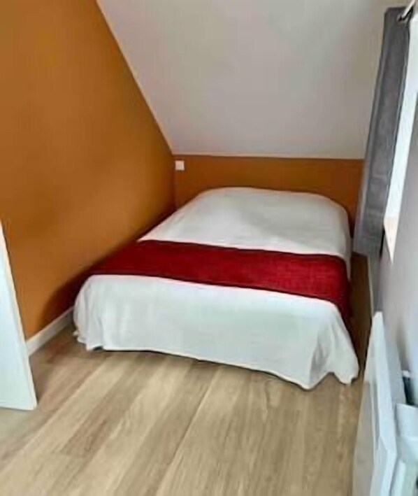Ein kleines Zimmer mit einem Einzelbett, geneigter Decke, Holzboden und einer roten Tagesdecke.