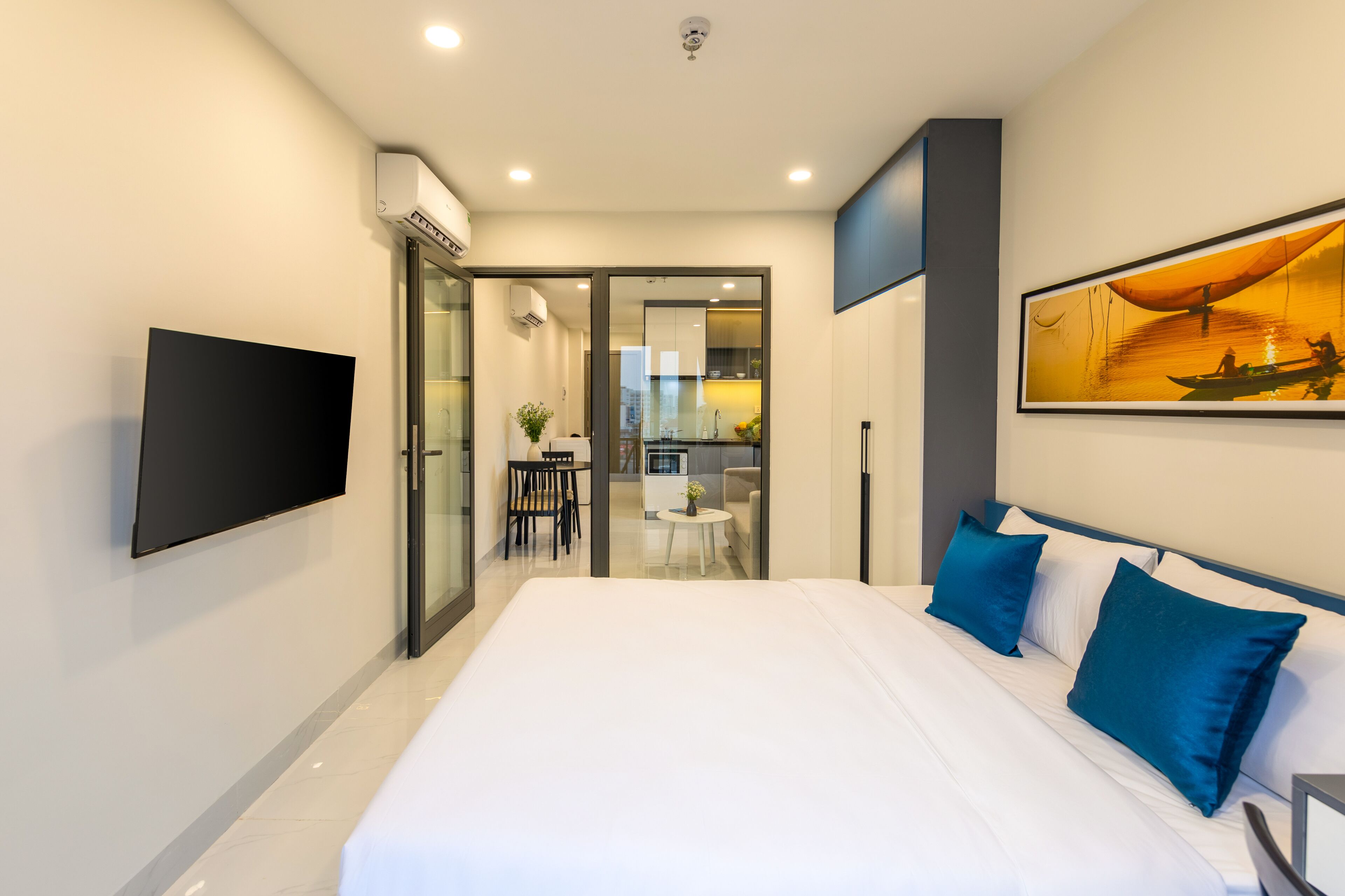 A modern hotel room with a large bed, a flat-screen TV, a painting, and a view into the kitchen area.