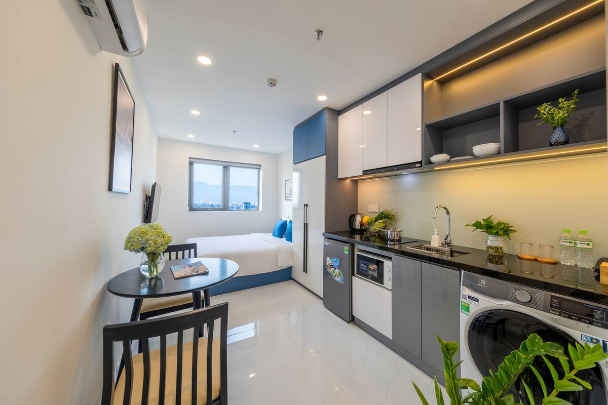 A compact kitchen with a washing machine, a dining area with a table and chairs, and a bed with a window view.