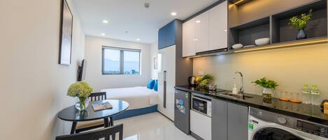 A compact kitchen with a washing machine, a dining area with a table and chairs, and a bed with a window view.