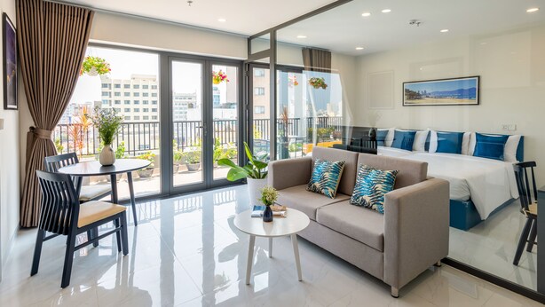 A modern hotel room with a sofa, a dining table, a small coffee table, and a bed with blue and white bedding. - The Home Hotel & Apartment (Da Nang)