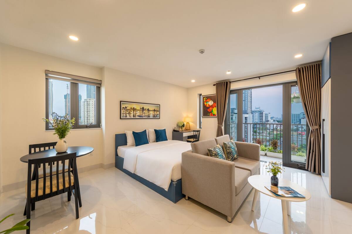 A modern hotel room with a large bed, a sofa, a dining table, and a view of the city.