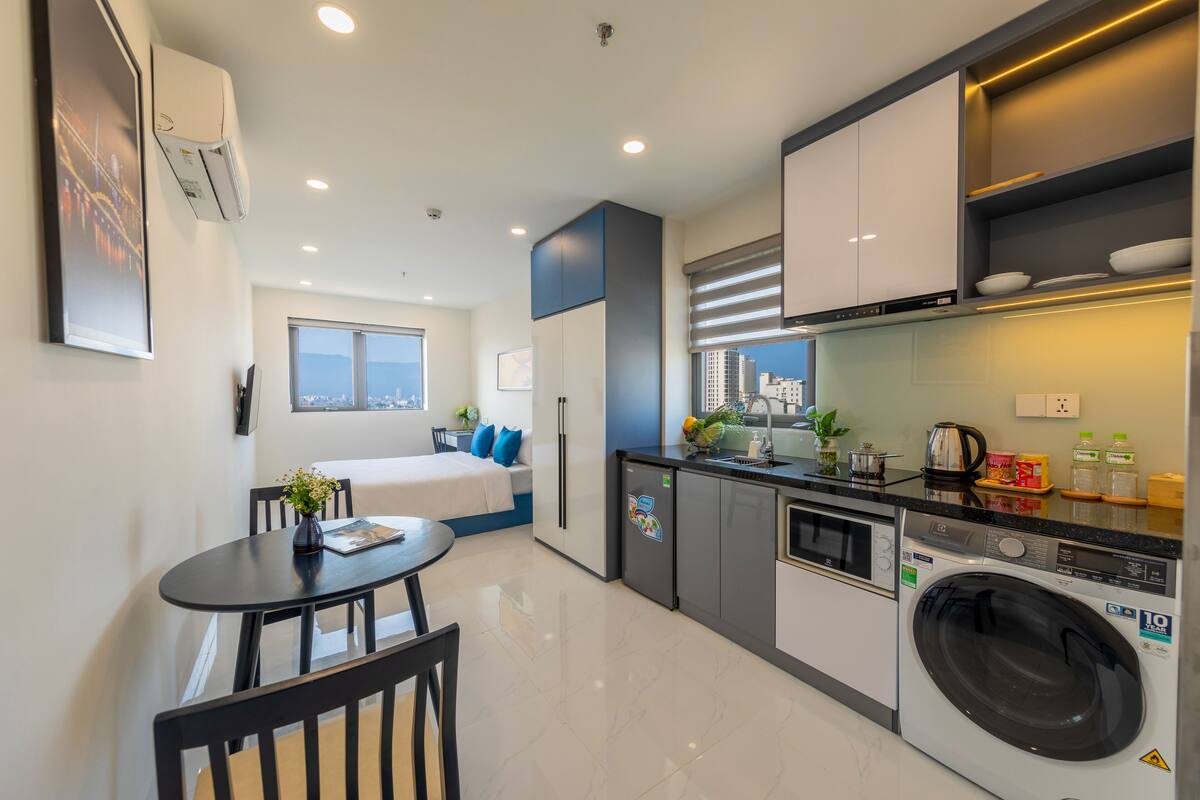 A modern kitchen with a built-in washer and dryer, a dining area with a table and chairs, and a bedroom with a bed and window.