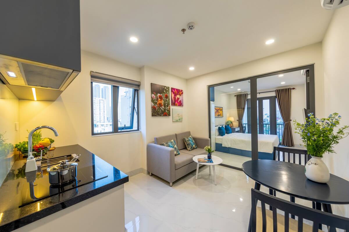 A modern kitchen with a sink, stove, and countertop. There is a gray sofa with a coffee table and a dining table with chairs. A window provides a view of the city.